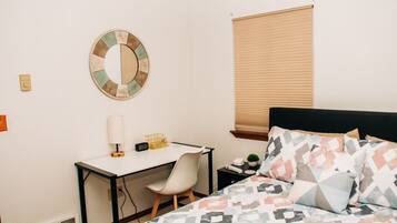 A bedroom with a bed, a desk, a chair, a lamp, a clock, a small plant, and a window with blinds.