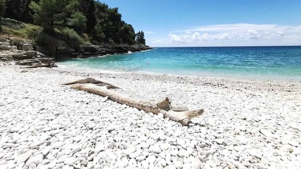 Une plage de galets bordée d’une eau turquoise cristalline et un promontoire rocheux parsemé d’arbres.