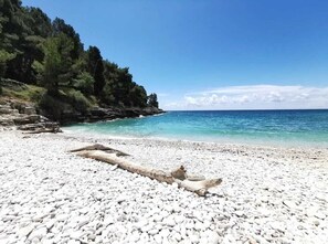 A pebble beach with clear turquoise water and a rocky outcrop with trees.