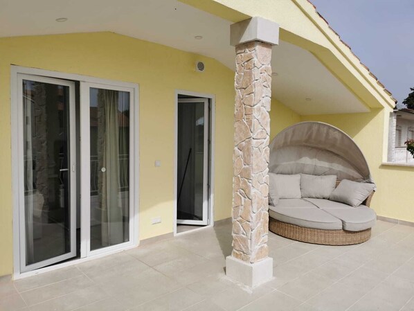 A covered outdoor seating area with a stone pillar, a glass door entrance, and a wicker sofa.