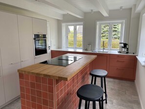 A modern kitchen with a central island, two bar stools, a built-in oven, and large windows.