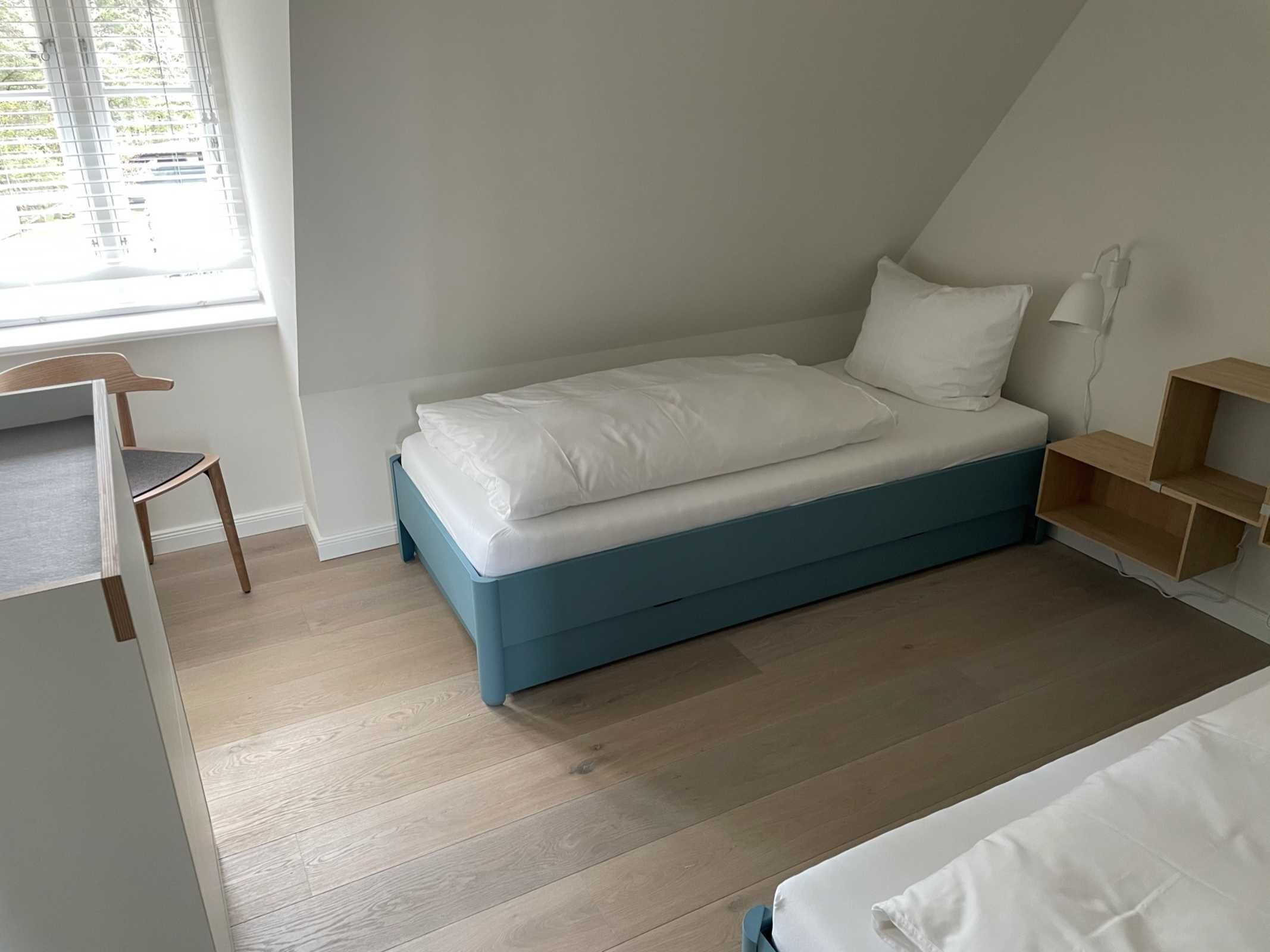 A small, single-bed room with a sloped ceiling, a wooden chair, a bedside table, and a window with blinds.