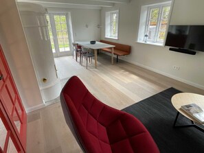 A modern living room with a red sofa, a dining table with chairs, a TV mounted on the wall, and large windows.