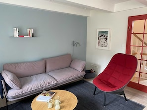 Una sala de estar moderna con un sofá, un sillón rojo, una mesa de centro con libros y velas, una estantería, una lámpara y un cuadro en la pared.