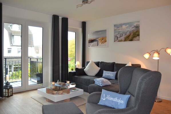 A modern living room with a grey sofa, a white coffee table, and a floor lamp. There are two windows with curtains, and a balcony is visible.