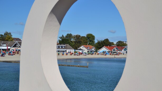 A view through a circular window of a waterfront area with buildings and a pier. - App. Ankerplatz mit 2 SZ und 2 Balkonen - Appartement mit 2 SZ (Kellenhusen (Ostsee))