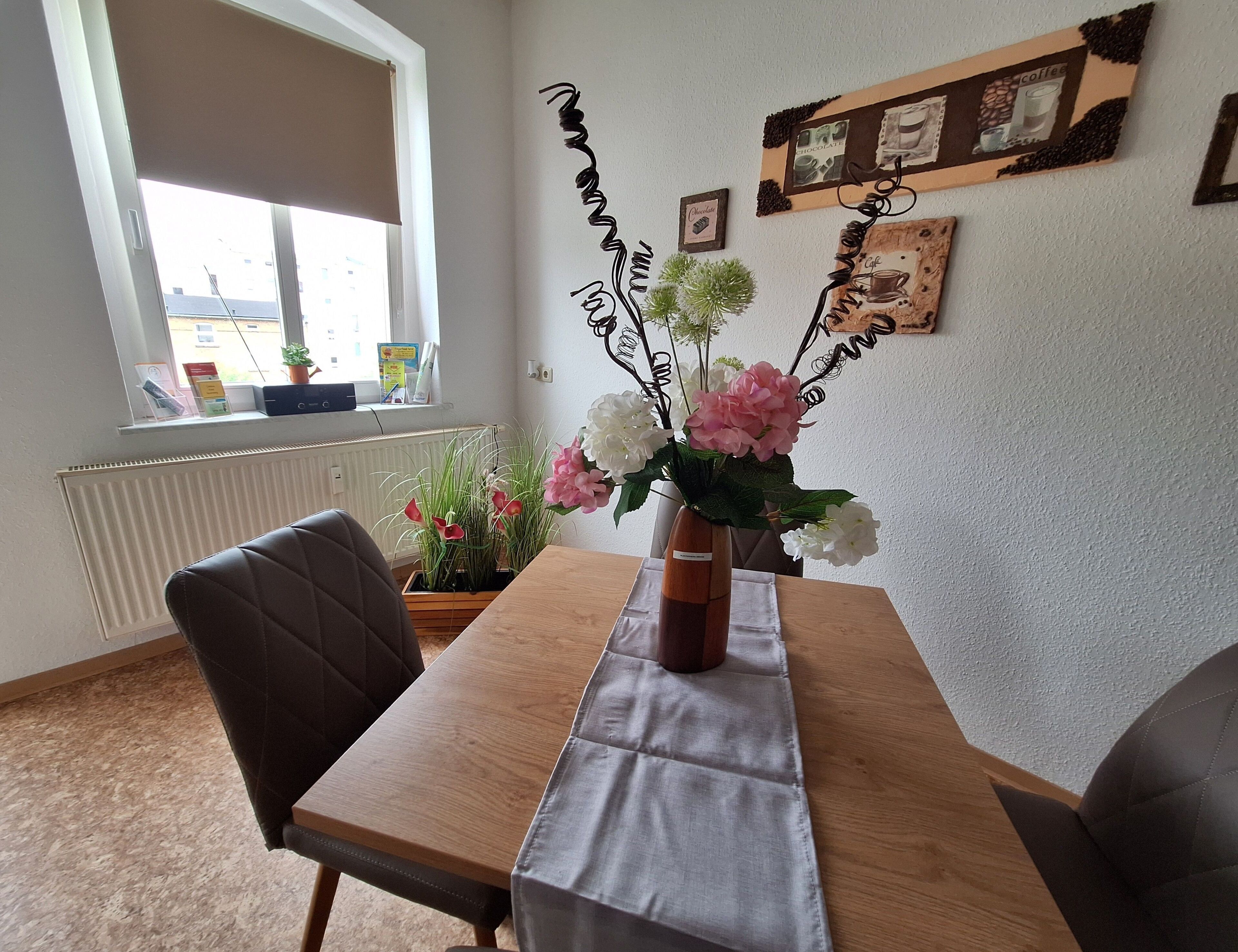 Ein Essbereich mit Tisch, Stühlen, einer Blumenvase, einem Fenster und Wanddekorationen.