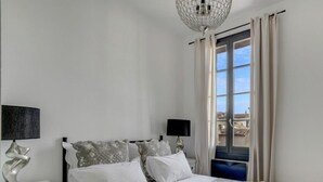 A neatly made bed with white linens and decorative pillows, a bedside table with a lamp, and a view of buildings through a window with curtains. - Beautiful Loft Apartement, quiet, 5 mn historical center (Isle sur la Sorgue)