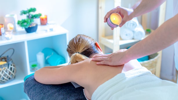 A massage therapy session in a spa setting with a client lying face down, a therapist applying a candle to their back, and a shelf with towels and candles in the background. - Morski Park Międzywodzie (Dziwnów)
