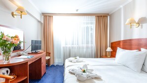 A hotel room with a wooden desk, a television, a bed with white linens, and a window with sheer curtains. - Morski Park Międzywodzie (Dziwnów)