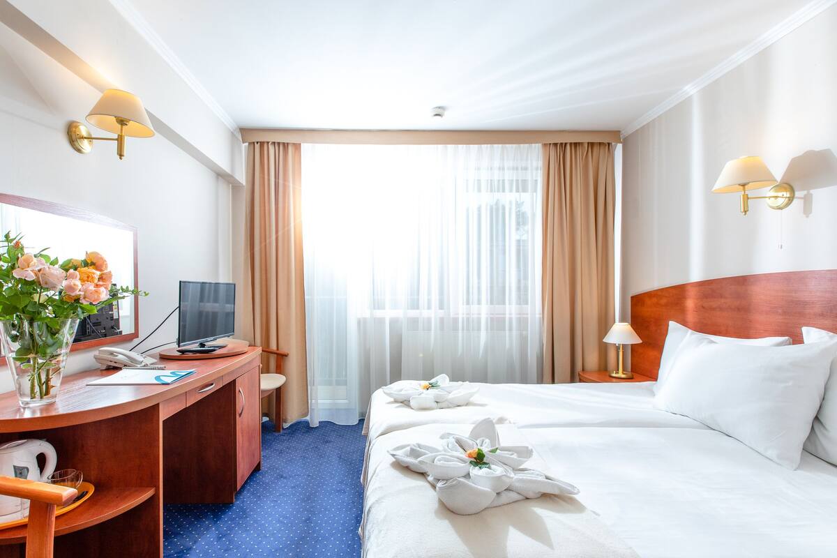 Hotellihuone, jossa on puinen työpöytä, televisio, valkoisilla lakanoiden päällä varustettu sänky ja ikkuna, jossa on läpikuultavat verhot.