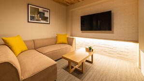 A modern living room with a beige sofa, wooden coffee table, and a flat-screen TV mounted on a white brick wall. - GRAND BASE Saiwaimachi (Nagasaki)