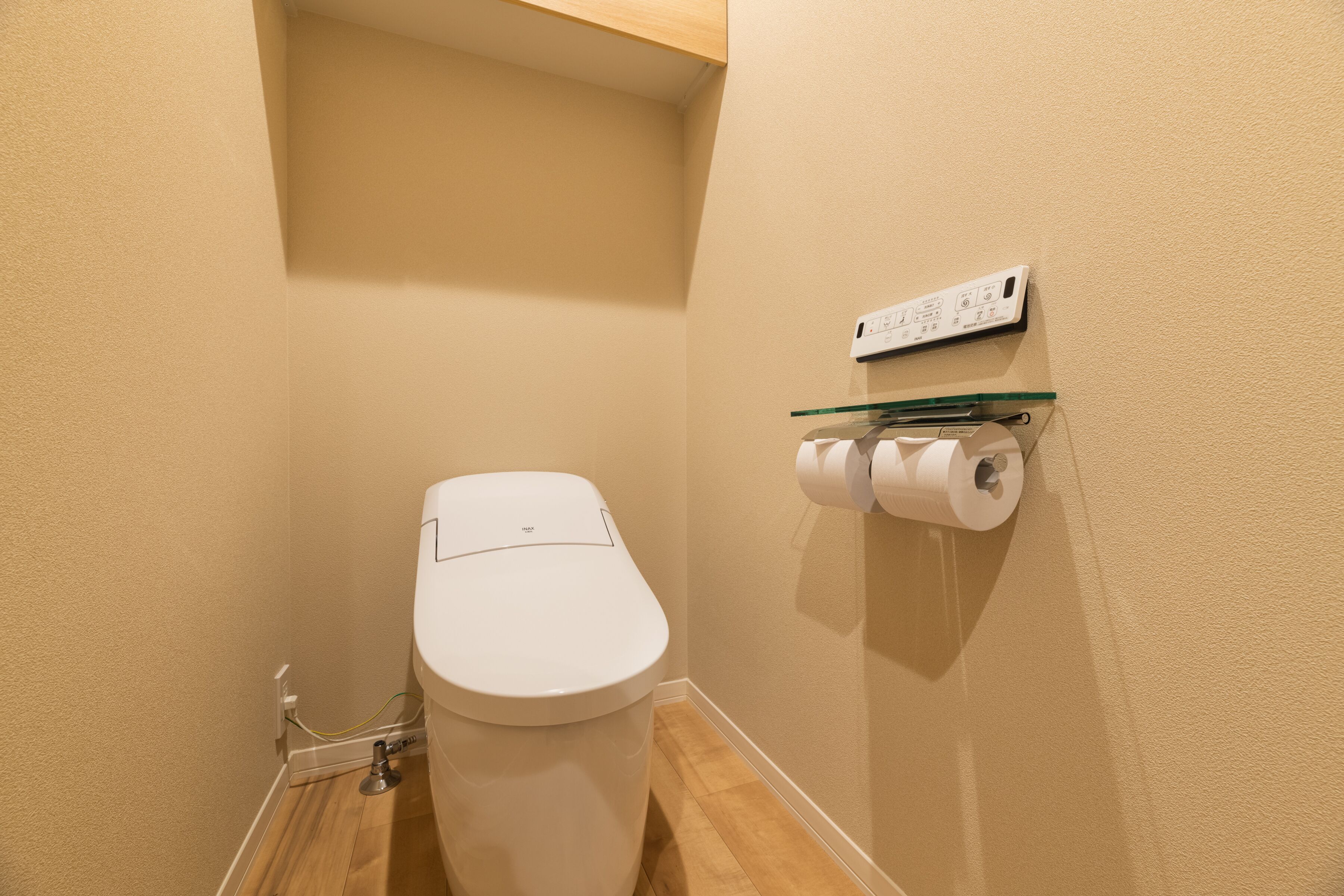 A small, clean bathroom with a white toilet, a glass shelf holding toilet paper, and a wall-mounted electrical outlet.