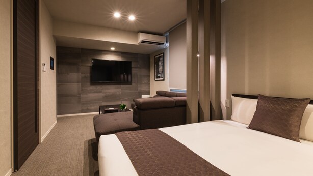 A modern hotel room with a large bed, a flat-screen TV mounted on the wall, a seating area with a sofa and ottoman, and a small table with a plant. - GRAND BASE Saiwaimachi (Nagasaki)
