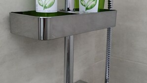 Une salle de bain équipée d’une pomme de douche chromée, d’un robinet chromé et d’une étagère où sont posés deux flacons de shampoing et d’après-shampoing « AMO ESSERE Eco ».