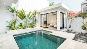 A modern pool area with a glass-enclosed lounge, surrounded by tropical plants and a white exterior.