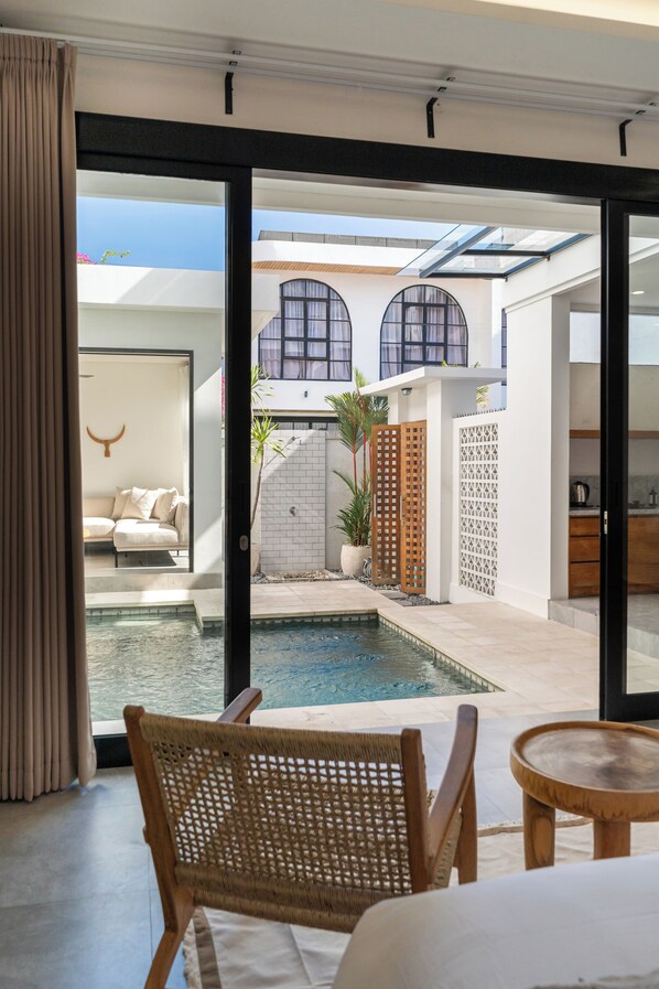 A modern hotel room with a large sliding glass door leading to a private outdoor area with a pool, a seating area with a sofa, and a dining table.