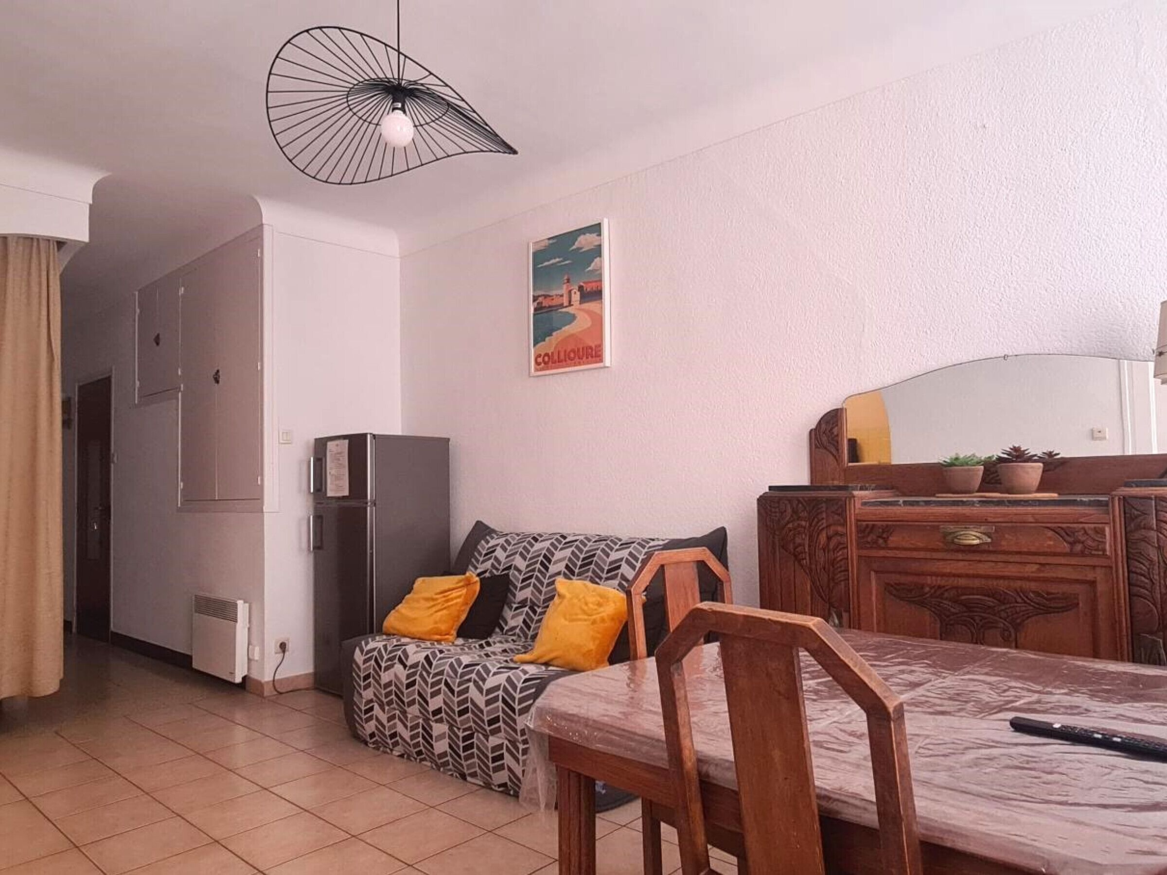 A compact living space with a sofa, a dining table with chairs, a refrigerator, a heater, and a decorative mirror.