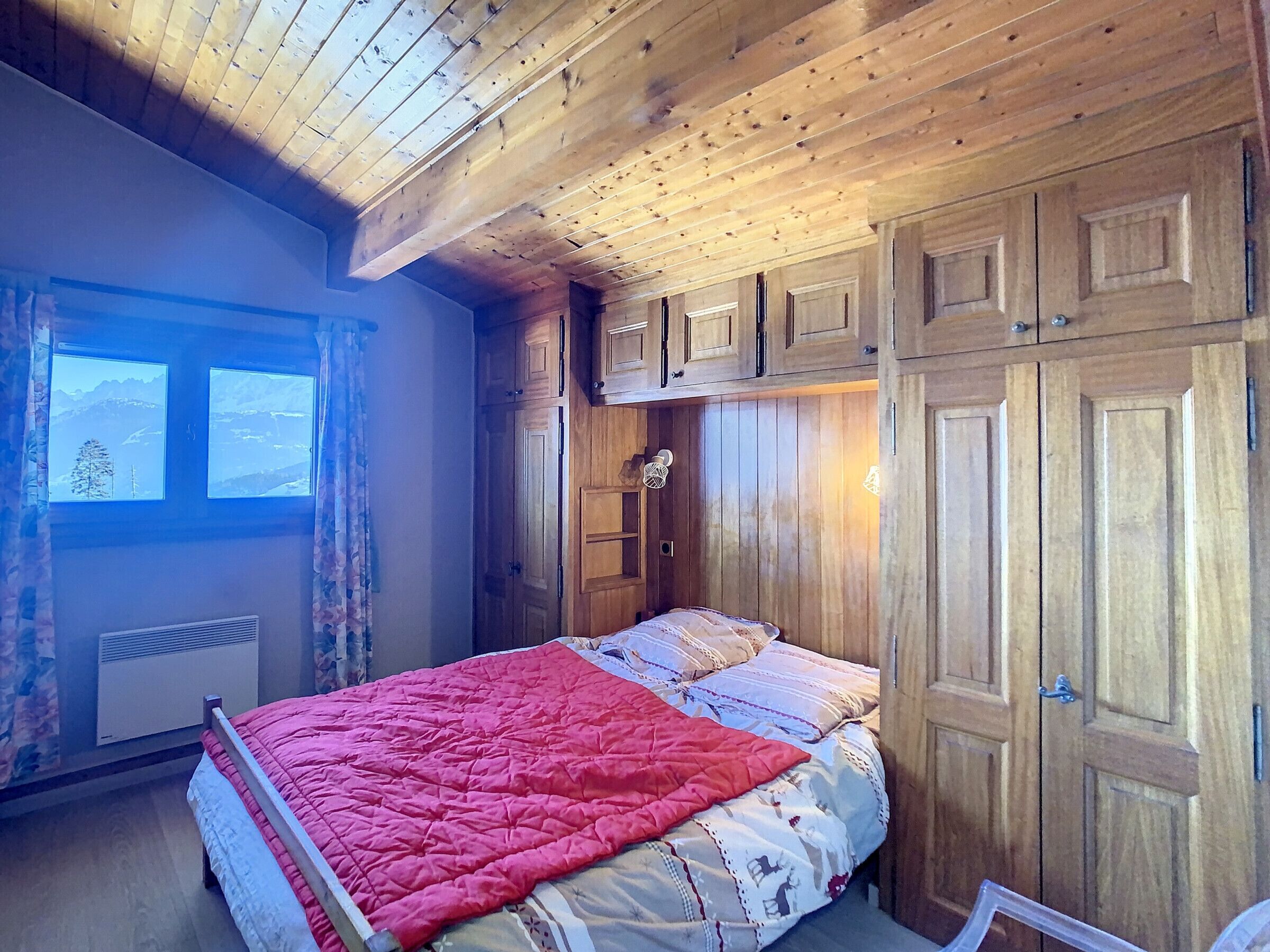 Ein Schlafzimmer mit Holzbalkendecke, einem Bett mit roter Decke, einem Fenster mit Bergblick und Holzschränken.