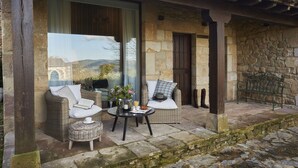 A stone building with a covered patio area featuring wicker furniture, a table with a vase and a book, and a view of the countryside.