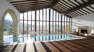 Una amplia piscina interior con cubierta de madera, grandes ventanales que ofrecen vistas al exterior y una chimenea.
