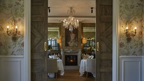 A formal dining room with a fireplace, two chandeliers, and floral wallpaper.