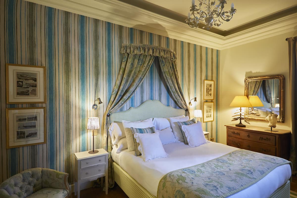 A neatly arranged bedroom with a four-poster bed, a bedside table with a lamp, a chair, and a dresser with a mirror.