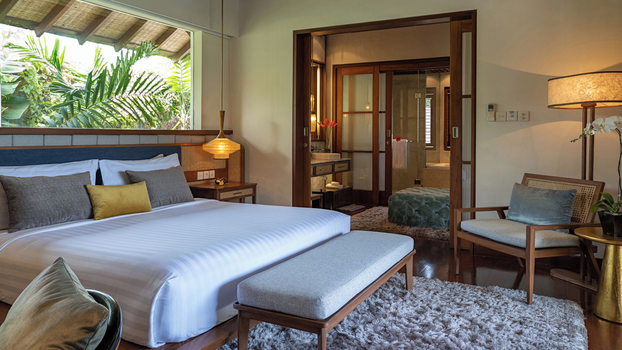 A spacious bedroom with a large bed, a sitting area with a chair and bench, a small table, and a view of a bathroom and a balcony with plants.