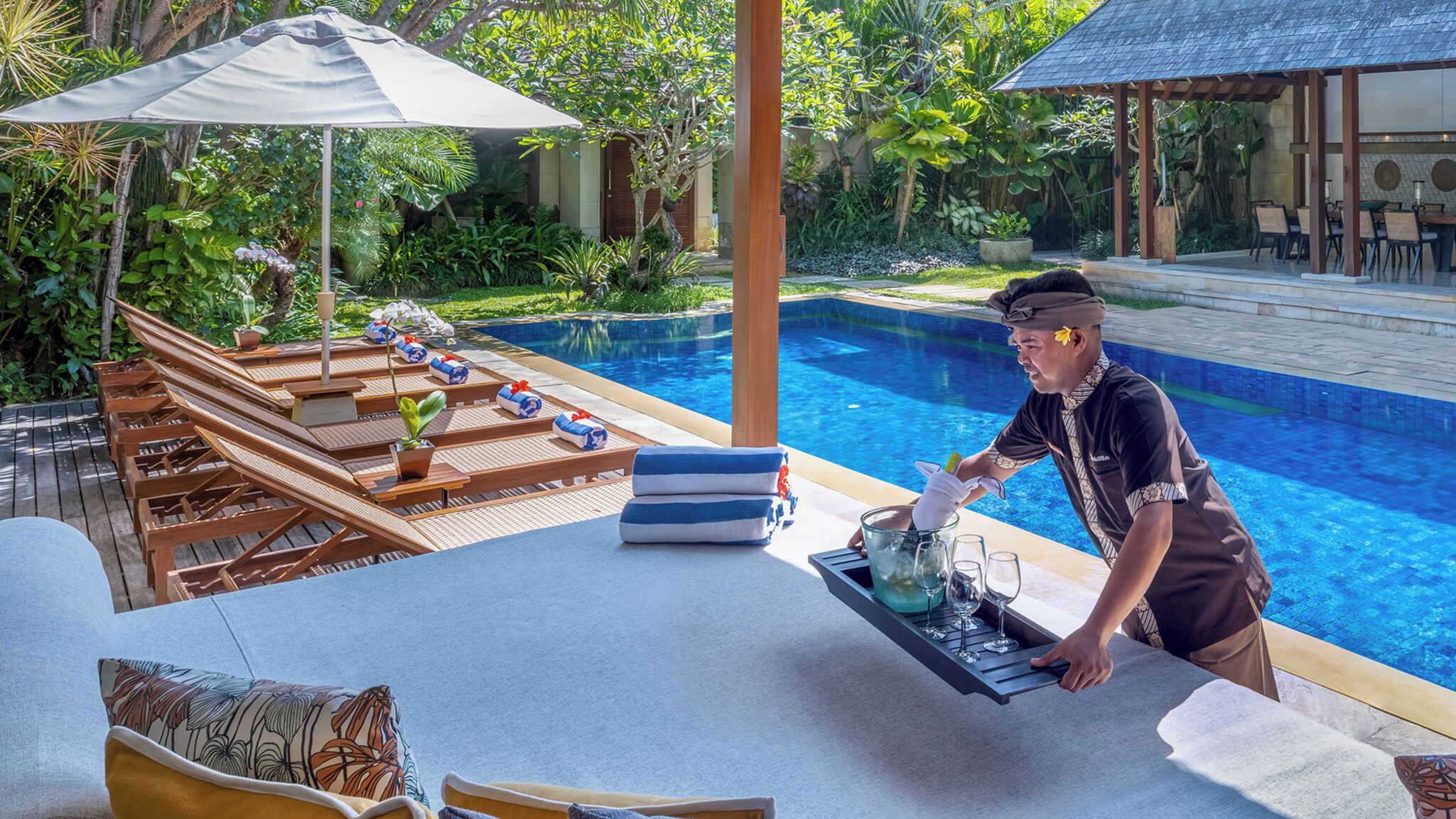 Un espace aménagé au bord de la piscine, avec des chaises longues, une table sur laquelle se trouve un plateau de verres et une bouteille, et un serveur qui s’affaire.