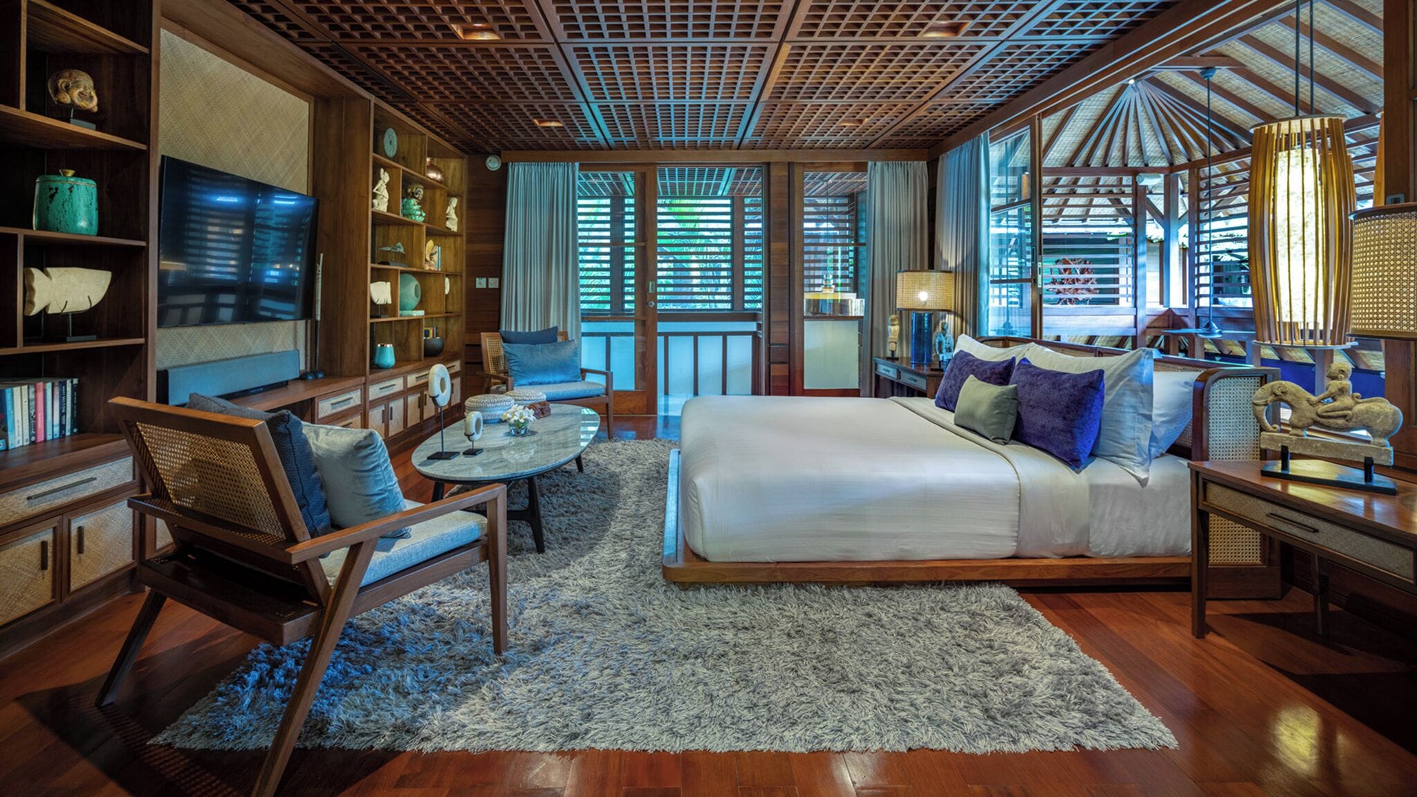 A spacious bedroom with a large bed, a sitting area with a sofa and chairs, a small round table, and a bookshelf filled with books and decorative items.