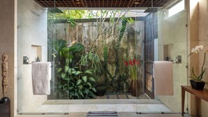 Een douchecabine met glazen wanden en uitzicht op een tropische tuin, met grote planten en een stenen muur.