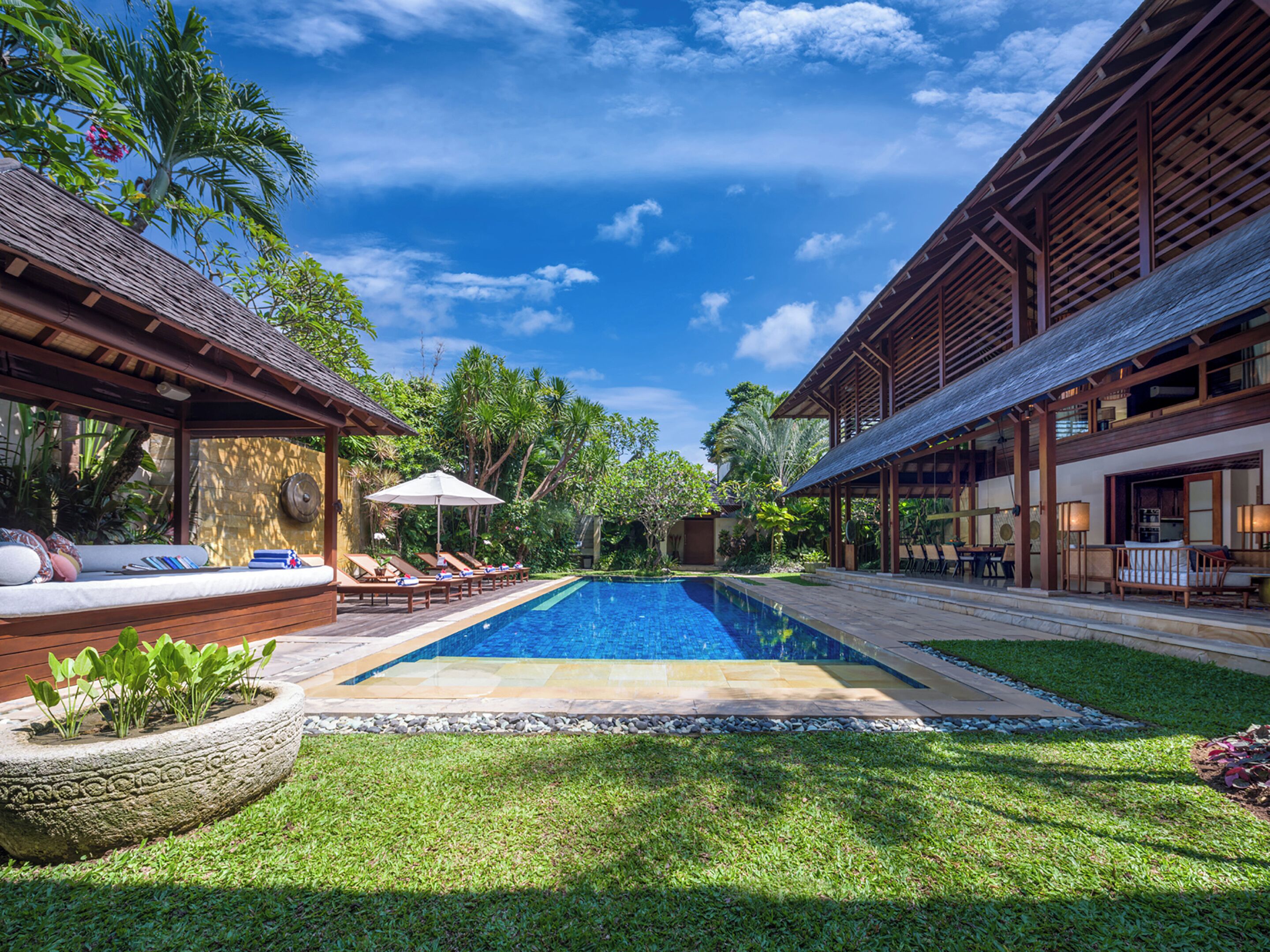A pool area with lounge chairs and a covered seating area, surrounded by lush greenery and a clear blue sky.