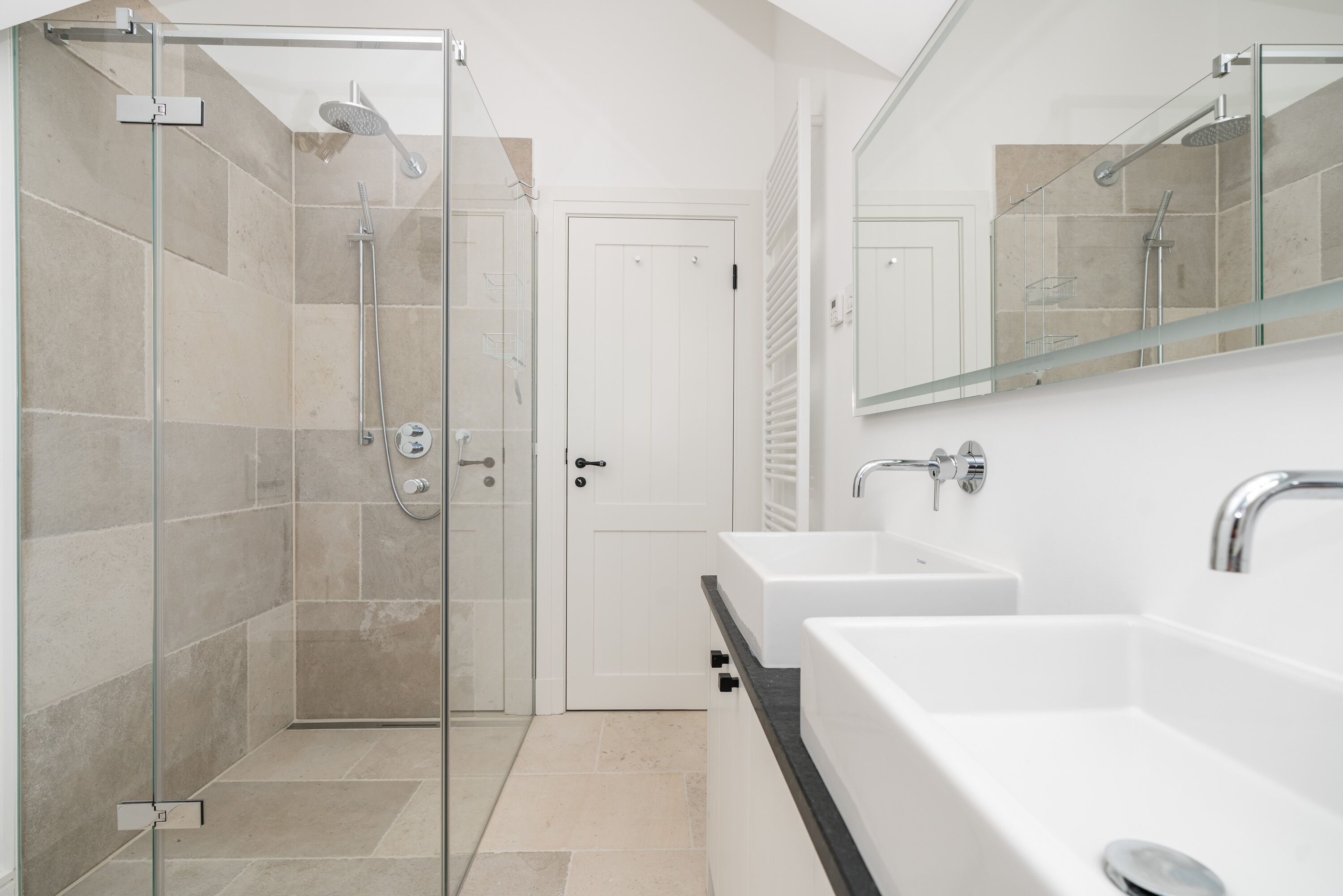 Une salle de bain moderne avec une douche à parois de verre, une porte blanche et un grand miroir au-dessus d’un lavabo blanc doté d’un plan de travail noir.