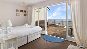 A bedroom with a bed, a nightstand, a lamp, a window with curtains, and a view of a balcony and beach. - Kilmore - Beach Front House Direct Beach Access Spectacular Sea Views (Saundersfoot)