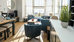 A modern living room with a wooden dining table, blue chairs, and a sofa. A TV is on the left, and there is a large window with blinds. - The Kings Vista (Weymouth)