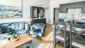 A modern kitchen with dark cabinets and a patterned backsplash, a dining area with blue upholstered chairs and a wooden table, and a living room with a blue sofa and a painting on the wall. - The Kings Vista (Weymouth)