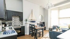 A modern kitchen with a large island, dark cabinets, and a patterned backsplash. There is a dining area with a table and chairs, and a view of the outside through large windows. - The Kings Vista (Weymouth)