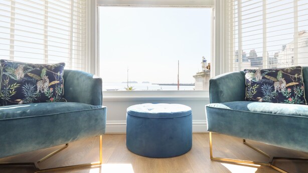 A room with two blue armchairs, a round ottoman, and a large window with blinds. - The Kings Vista (Weymouth)