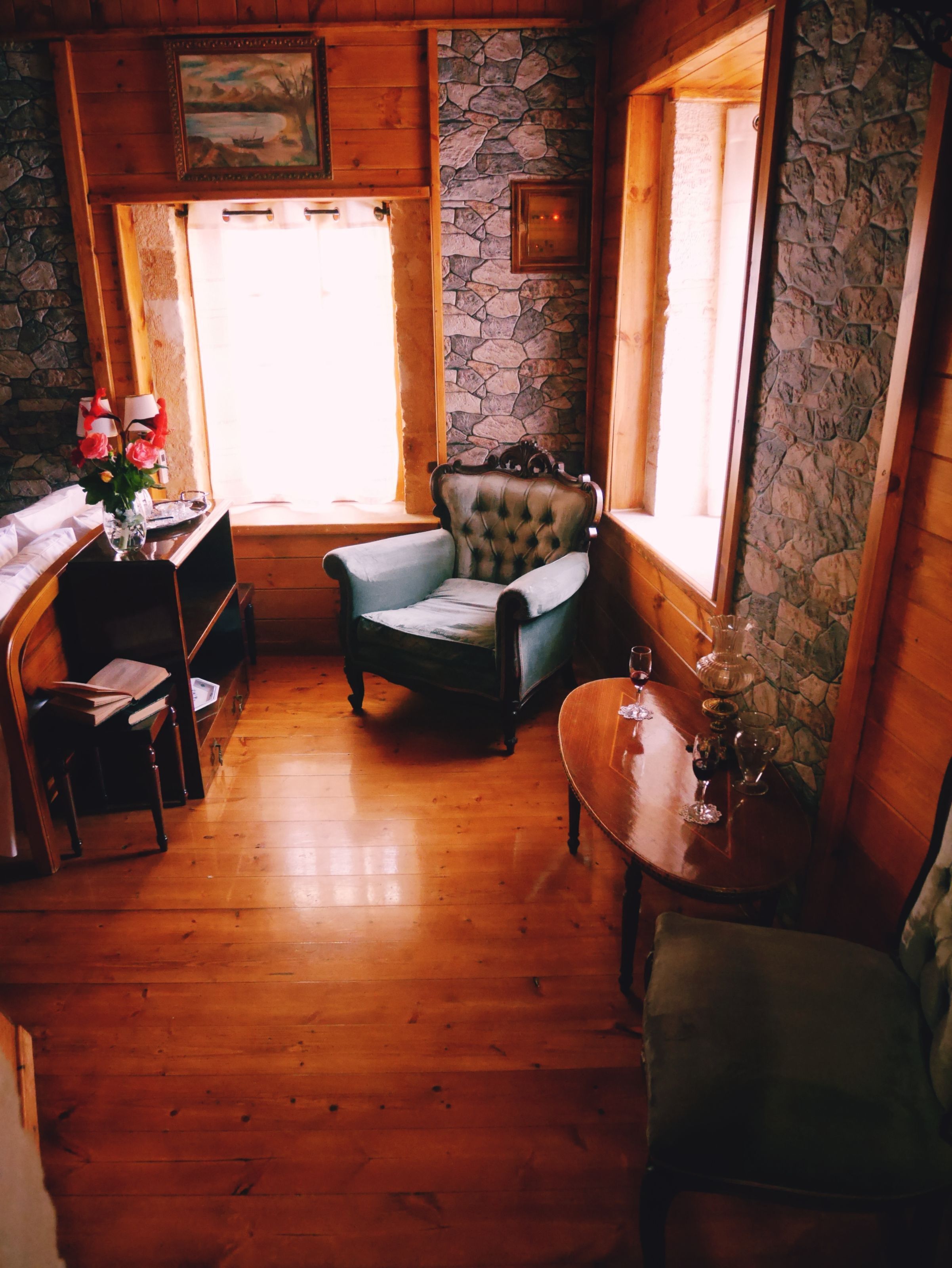 A cozy room with wooden floors, a stone wall, a cushioned armchair, a wooden table with a vase, and a desk with a lamp.