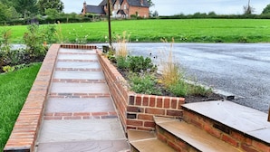 Un chemin pavé de briques mène à un escalier également en briques, avec un champ herbeux et une maison en arrière-plan.