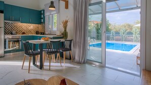 Una cocina moderna con gabinetes color turquesa, un comedor con mesa y sillas, y un amplio ventanal corredizo que da a una piscina.