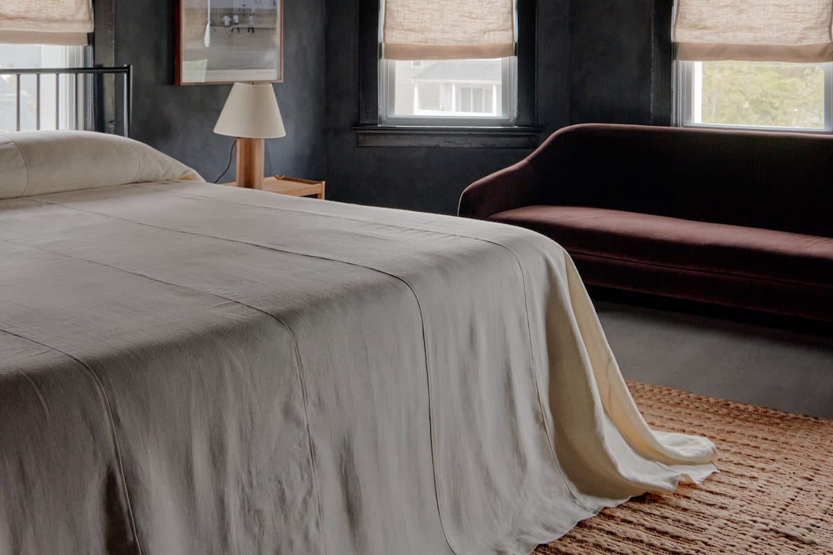 Een slaapkamer met een bed, een bankje, een lamp en twee ramen met jaloezieën.
