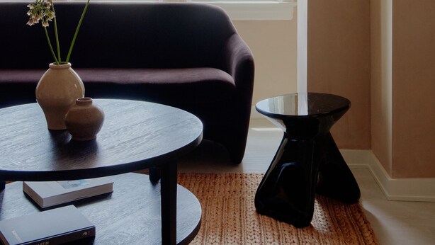 Un salon avec un canapé foncé, une table basse ronde, une petite table d’appoint et un vase rempli de fleurs.