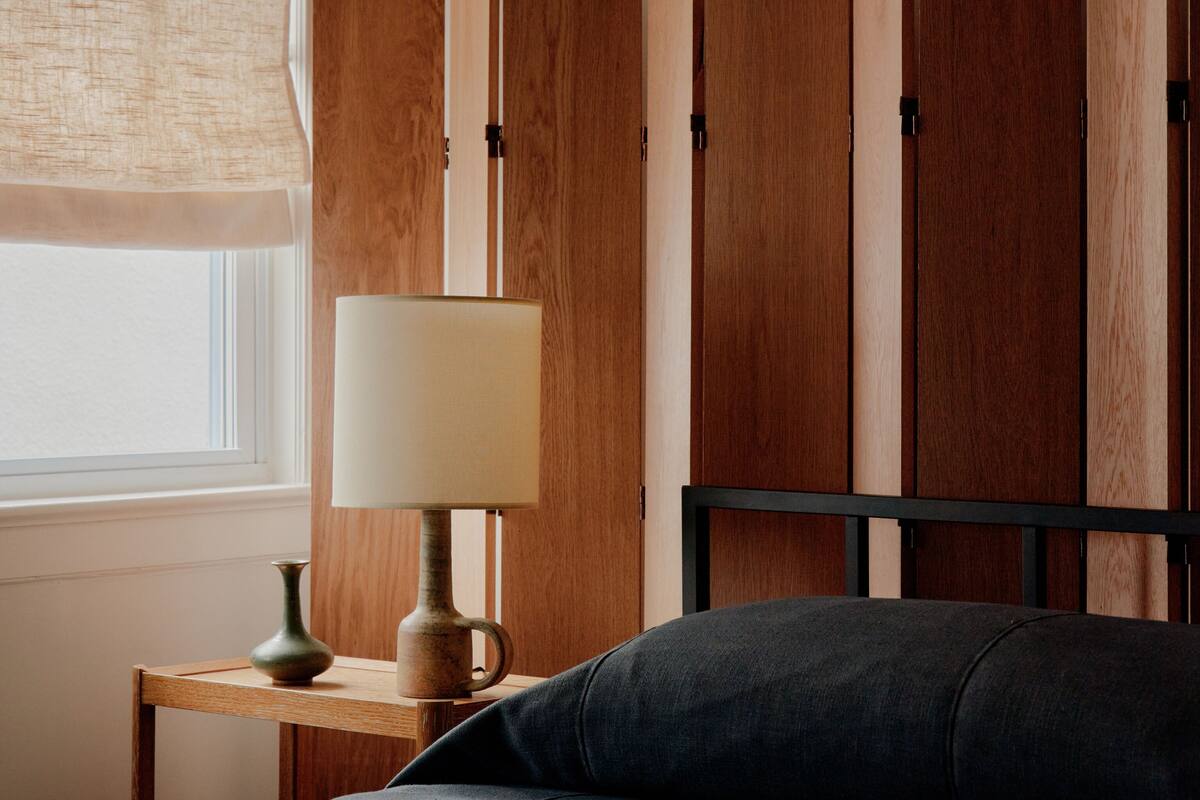 Een slaapkamer met een houten garderobe, een nachtkastje met een lamp, en een bed met een grijs matras.