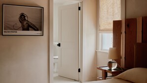 Une chambre d’hôtel avec un lit, une table de chevet avec une lampe, un tableau encadré au mur et une porte donnant sur une autre pièce.