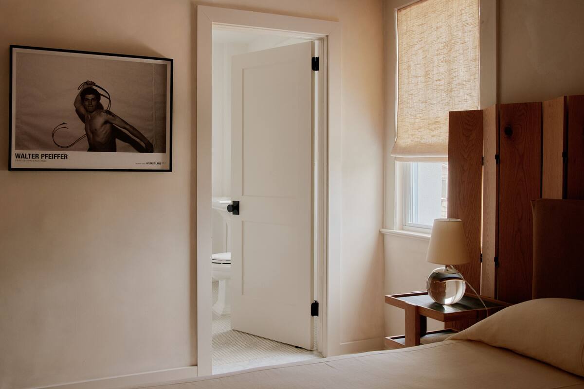 Een hotelkamer met een bed, een nachtkastje met een lamp, een lijst aan de muur, en een deur naar een andere kamer.