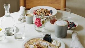 Une table à manger avec des aliments pour le petit-déjeuner, une carafe, des verres et un vase rempli de fleurs.
