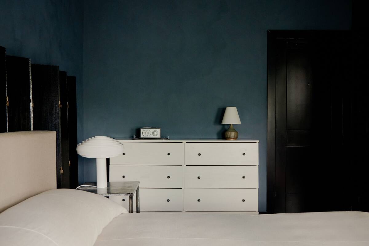 Een slaapkamer met een bed, een dressoir, een nachtkastje, een lamp en een klok.