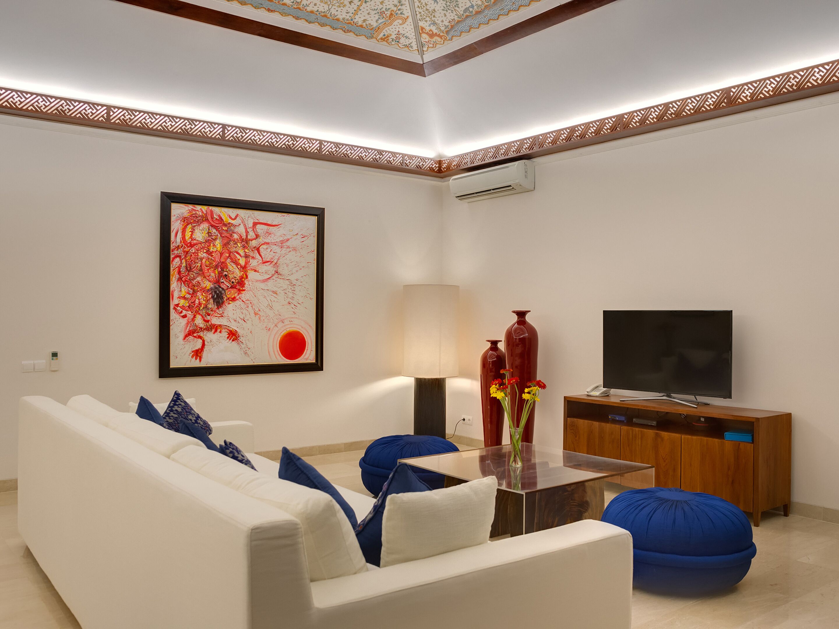 A modern living room with a white sofa, blue cushions, a glass coffee table, a red vase with flowers, a flat-screen TV, and a wooden cabinet.