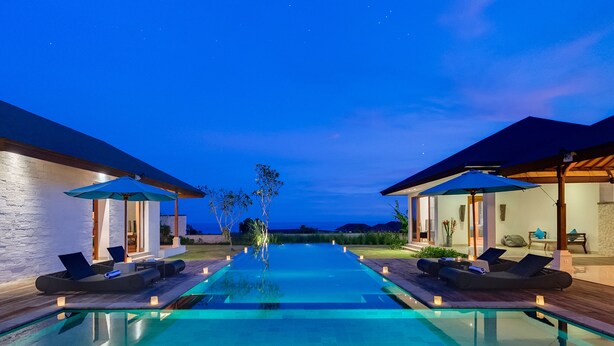 A modern infinity pool with lounge chairs and umbrellas, surrounded by well-lit buildings with large windows. - Pandawa Cliff Estate (Kutuh)